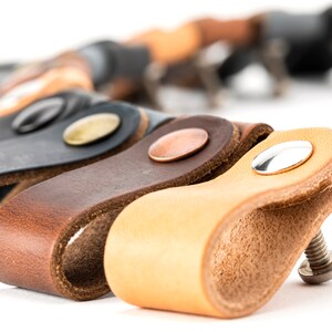 May include: Close-up of several leather drawer pulls in various colors, including tan, brown, and gray. Each pull features a loop design with a metal rivet and a screw for installation. The pulls are arranged in a row against a white background.