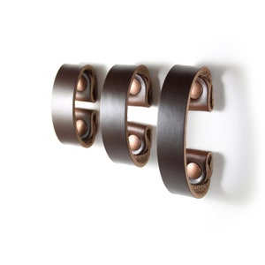 May include: Three brown leather hooks with copper rivets. The hooks are mounted on a white wall.