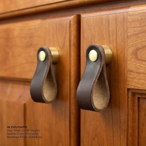 May include: Two cabinet pulls with a chocolate brown leather loop and solid brass hardware. The pulls are attached to a wooden cabinet door. The height of the pulls is 2-1/4 inches.