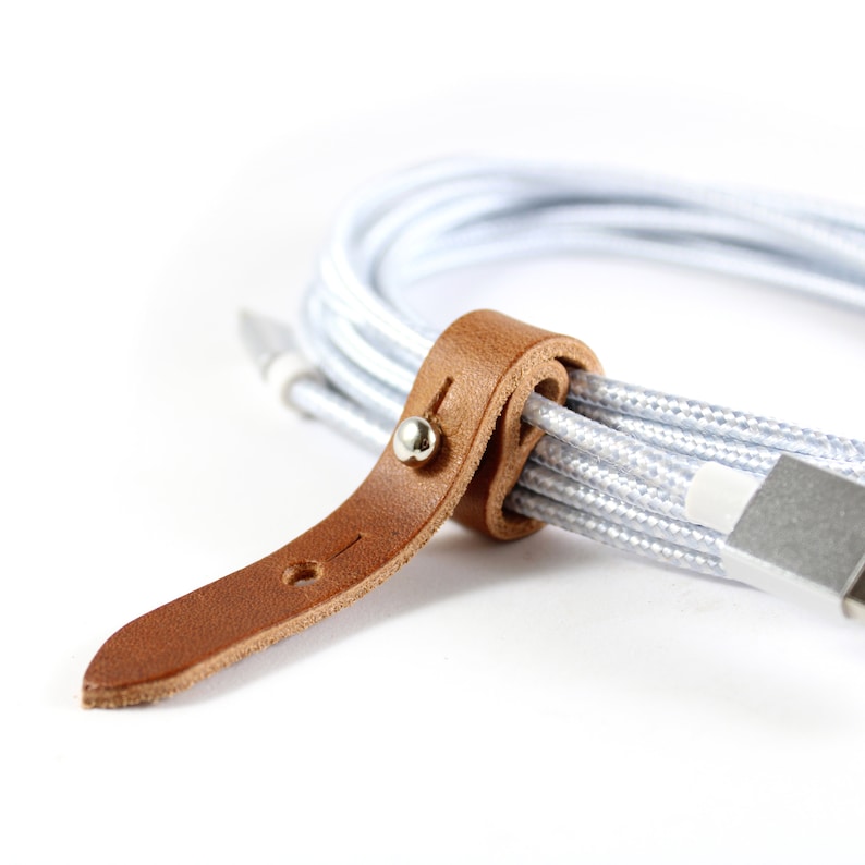 May include: A brown leather cable organizer with a silver metal button. The organizer is wrapped around a white cable.