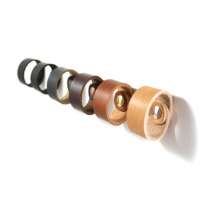 May include: A set of eight leather rings in various shades of brown, black, and tan. The rings are arranged in a line, with the darkest ring on the left and the lightest ring on the right. Each ring has a brass rivet in the center.