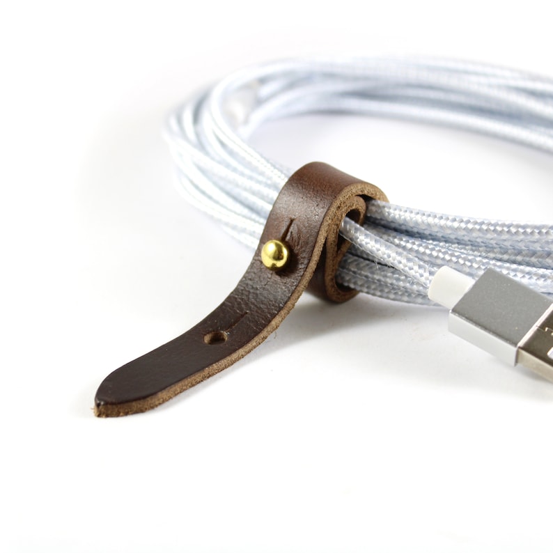 May include: A brown leather cord organizer with a gold metal button. The organizer is wrapped around a white cable.