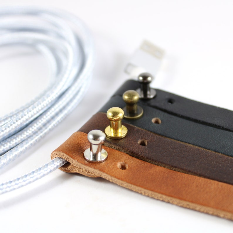 May include: A close-up of a leather cord organizer with four different colored leather straps and metal snaps. The straps are brown, dark brown, black, and gray. The snaps are silver, gold, and brass.