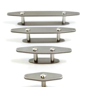 May include: A series of four metal cabinet pulls, each with a brushed silver finish. The pulls have an oval-shaped top with two small, rounded silver rivets and a pair of cylindrical legs. The pulls are arranged vertically against a white background.