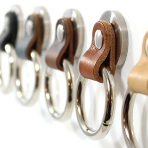 May include: Five silver key rings with leather loops. The loops are in various shades of brown. The rings are attached to a white wall.