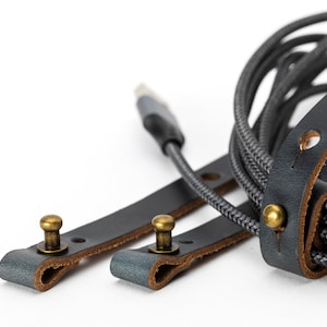 May include: Two black leather straps with brass buckles and a gray braided cord. The straps are designed to organize and secure cords.