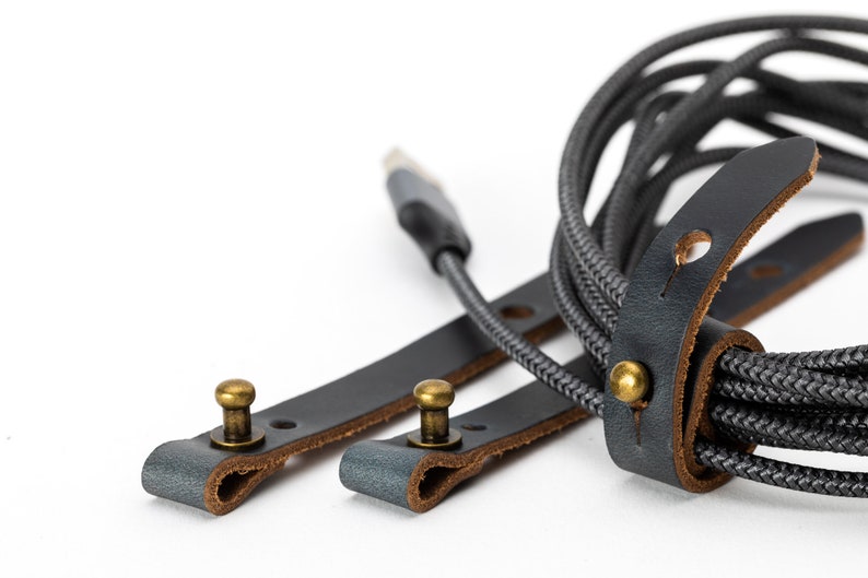 May include: Two black leather straps with brass buckles and a gray braided cord. The straps are designed to organize and secure cords.