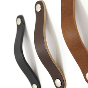 May include: Three curved leather drawer pulls in black, dark brown, and light tan. Each strap features a smooth finish and is secured at both ends by a circular silver-toned metal rivet. The handles are arranged vertically in a row.