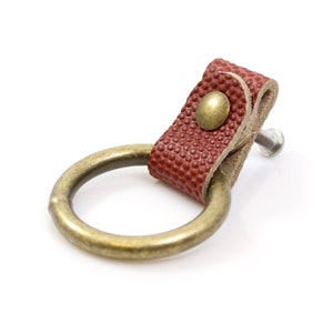 May include: A red leather drawer pull with a brass ring and a brass screw. The leather has a textured pattern.