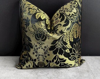 May include: Cushion with a black and gold floral design. The pillow has a black border and is placed on a grey surface. The pattern features stylized flowers and leaves in a repeating design, creating a luxurious aesthetic.