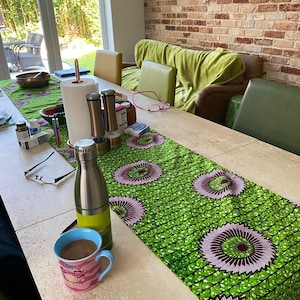 May include: A dining table features a green and purple patterned table runner. A stainless steel water bottle and a pink and blue mug with a cartoon design are on the table. Paper towels and spice grinders are also visible on the table.