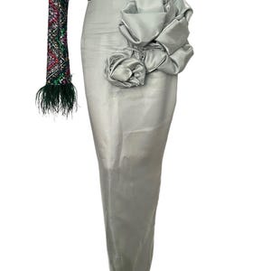 May include: A silver-gray formal gown with a large floral embellishment. The bodice is adorned with colorful sequins, and one long sleeve features a feather trim. The skirt is a sleek, floor-length design.