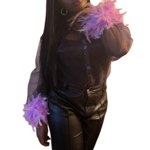 May include: A woman wearing a black sheer blouse with purple feather trim on the sleeves and black leather pants.
