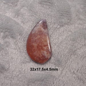 May include: A polished, teardrop-shaped gemstone with a mix of red, orange, and white hues. The stone has a smooth, glossy surface and measures 1.26 x 0.69 x 0.18 inches. The gemstone is set against a soft, gray background.