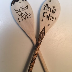 May include: Two wooden spoons with Harry Potter themed designs. One spoon has the text "The Boy Who Lived" with a lightning bolt. The other spoon has the text "Accio Cake" with a star design.