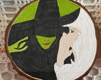 Puede incluir: Adorno redondo de madera con un diseño dividido de dos personajes. Un lado representa una figura de piel verde con un sombrero negro, el otro una figura de piel clara. El adorno está pintado con acrílicos y tiene un pequeño lazo metálico para colgar.