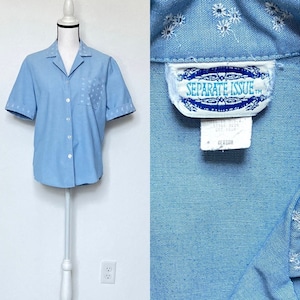 Puede incluir: Una camisa de manga corta azul claro con un estampado floral blanco bordado. La camisa tiene cuello y un bolsillo en el lado izquierdo. La etiqueta dice "Separate Issue".