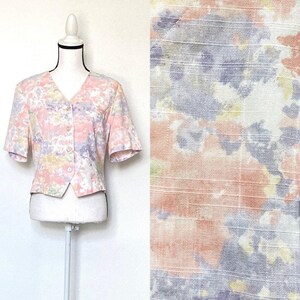 May include: A short-sleeved, button-up blouse with a pink, yellow, and purple abstract print. The blouse has a v-neck and a cropped fit.