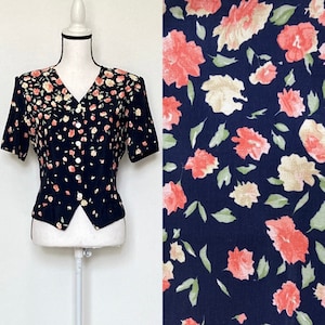 May include: A navy blue short-sleeved blouse with a floral print featuring pink and white flowers. The blouse has a v-neck and button closure.