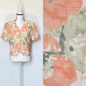Può includere: Una camicetta a maniche corte con bottoni con un motivo floreale nei toni del pesca, verde e crema. La camicetta è esposta su un manichino bianco. Il tessuto ha un aspetto strutturato. Il motivo floreale è mostrato anche in primo piano.