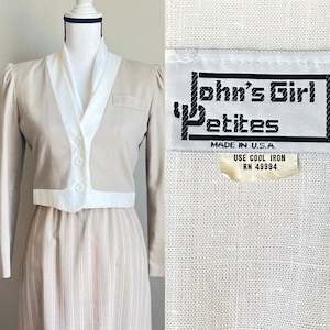 Może przedstawiać: Spódnica w pasy beżowe i białe oraz beżowa, krótka kurtka z białym kołnierzem i lamówką. Kurtka ma kieszeń po lewej stronie i trzy guziki z przodu. Etykieta na kurtce głosi "John's Girl Petites" i "Made in U.S.A. Use Cool Iron RN 49994".