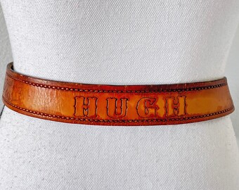 May include: A brown leather belt with the name "HUGH" embossed in white lettering.
