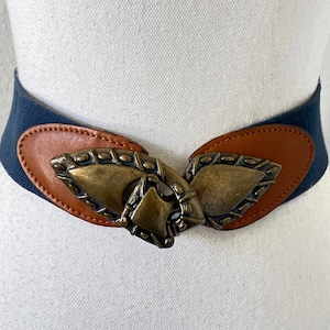 May include: A dark blue elastic belt with a brown leather trim and a large, ornate gold-toned buckle. The buckle is shaped like two overlapping leaves with intricate details.