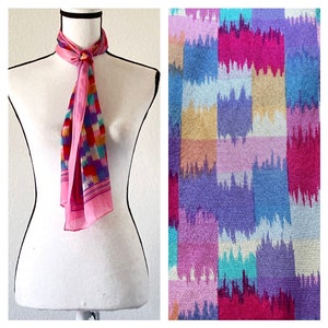 May include: A pink silk scarf with a colorful geometric pattern. The scarf is tied around a white mannequin's neck.