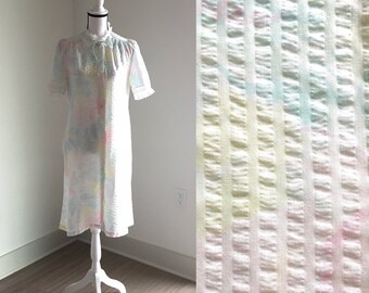 Puede incluir: Un camisón blanco con un estampado a cuadros en tonos pastel de rosa, azul y amarillo. El vestido tiene un cuello con volantes y mangas cortas.