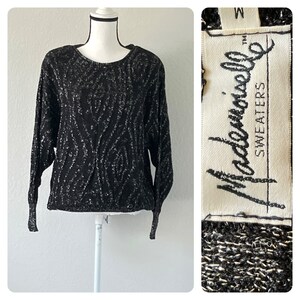 May include: Black and silver sweater with a sparkly, textured design. The sweater has a round neckline and long sleeves. The label reads "Madamemoiselle Sweaters".