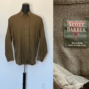 Puede incluir: Una camisa de manga larga de color verde oliva con botones y un bolsillo en el pecho. La camisa está hecha de 100% algodón y tiene una etiqueta que dice "Scott Barber XL 100% Algodón Hecho en Hong Kong".