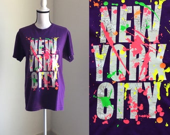 May include: Purple t-shirt with a white "NEW YORK CITY" graphic. The graphic has neon green, pink, and orange paint splatters.