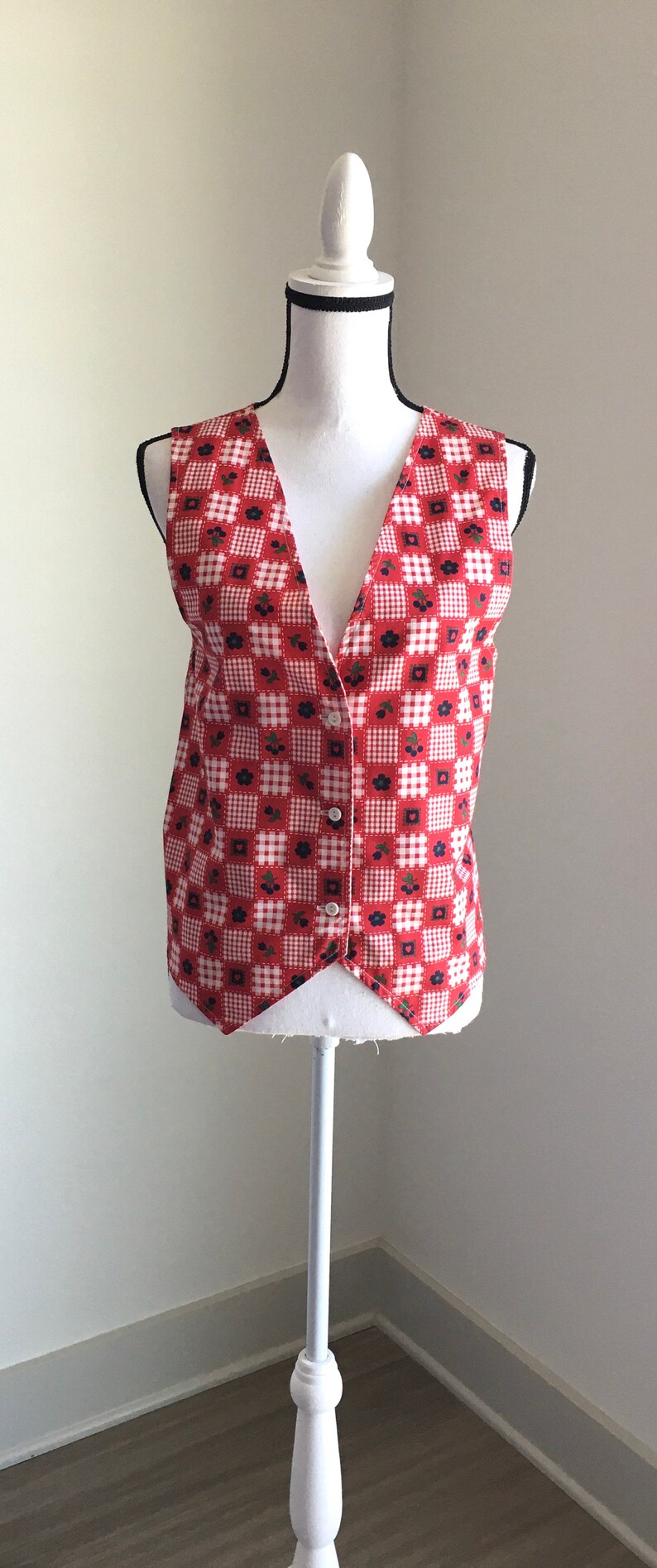 May include: A red and white gingham vest with a floral pattern and a button closure. The vest is sleeveless and has a V-neck.