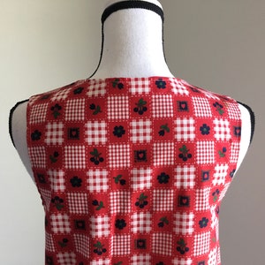 May include: Red and white gingham sleeveless vest with a patchwork design featuring small red hearts, black flowers, and green leaves with black berries.