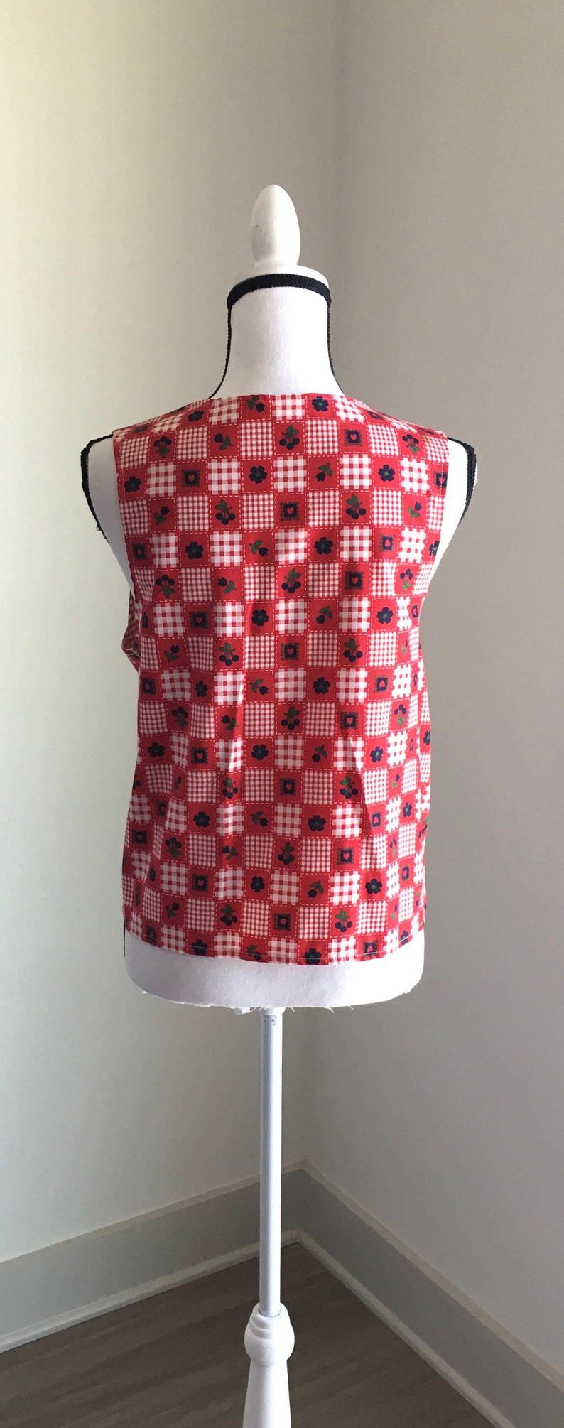 May include: A sleeveless red and white gingham vest with a floral pattern. The vest is on a white mannequin.