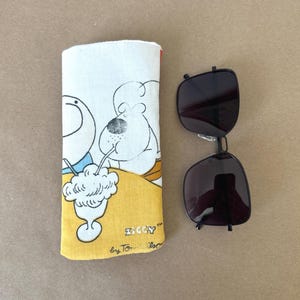 May include: A rectangular fabric eyeglass case featuring a cartoon illustration of Ziggy drinking from a milkshake with the text Ziggy by Tom Wilson, positioned next to a pair of black-framed sunglasses with dark lenses and a thin metal bridge.