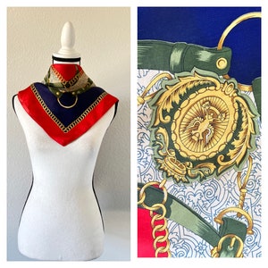 May include: A red, blue, and gold silk scarf with a chain pattern and a large gold emblem featuring a stylized bird. The scarf is displayed on a white mannequin.