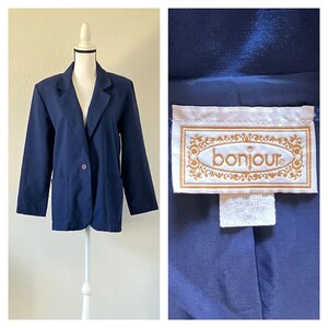 Puede incluir: Una chaqueta azul marino con un cierre de botón único y dos bolsillos. La chaqueta tiene una etiqueta que dice "bonjour" en letras doradas.