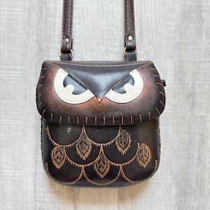 Könnte beinhalten: Braune Leder-Umhängetasche in Eulenform. Die Tasche hat ein detailliertes Eulengesicht mit weißen und braunen Akzenten und ein dekoratives Blättermuster. Sie hat einen braunen Riemen und ist zum Umhängen konzipiert.