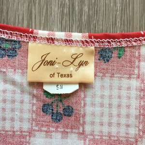 May include: A close-up of a white and red gingham fabric with a brown tag that reads "Joni-Lyn of Texas" and a white tag that reads "S M".