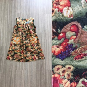 Puede incluir: Un vestido sin mangas con estampado floral verde, marrón y naranja que representa una cornucopia de frutas y verduras. El vestido tiene dos bolsillos en la parte delantera y un escote fruncido.