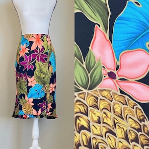 以下が含まれることがあります： ピンク、ブルー、グリーン、イエローのパイナップル、花、葉のトロピカルプリントが施された黒いスカート。スカートはフリルのあるヘムラインです。