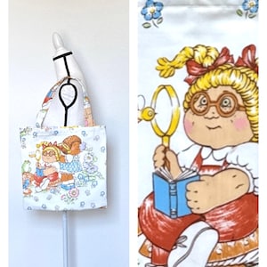 May include: A white fabric tote bag featuring a repeating pattern of cartoon characters with blonde hair, glasses, and red overalls, holding magnifying glasses and blue books. The design includes small blue flowers, green frogs, and birds.