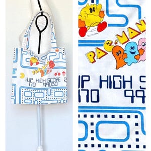 May include: White tote bag featuring a Pac-Man design with blue, yellow, orange, and pink characters. The bag includes the text "1UP HIGH SCORE 170 99030" and a blue maze pattern. The bag has a rectangular shape and two handles.
