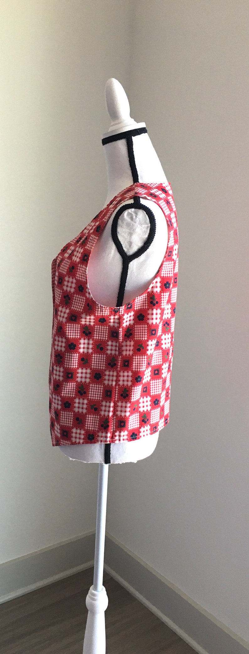 May include: A red and white gingham sleeveless vest with a floral pattern. The vest is on a white mannequin.