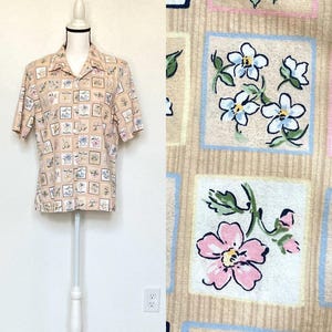 Puede incluir: Camisa de manga corta de color beige claro, adornada con un patrón floral repetido dentro de marcos cuadrados. Los diseños florales presentan flores rosas, azules y blancas. La camisa se exhibe en un maniquí blanco.