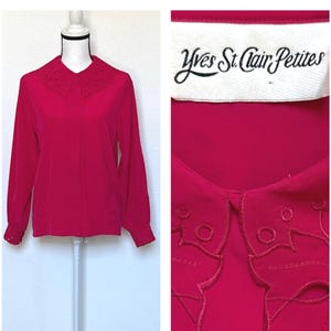 Könnte beinhalten: Eine langärmlige, fuchsiafarbene Bluse mit dekorativem Spitzenkragen und Manschetten. Die Bluse wird auf einer Schaufensterpuppe präsentiert. Das Etikett lautet "Yves St. Clair Petites".