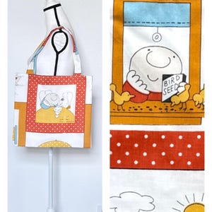 May include: A fabric tote bag featuring a cartoon illustration of a character in a window feeding yellow birds from a box labeled BIRD SEEDS. The bag has a white background with red polka dot borders, yellow accents, and a sun and cloud print.