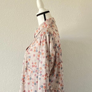 Puede incluir: Una camisa beige de manga larga con estampado floral de peque&ntilde;as flores rojas, azules y naranjas. La camisa est&aacute; en un maniqu&iacute; blanco con una banda negra en el cuello.