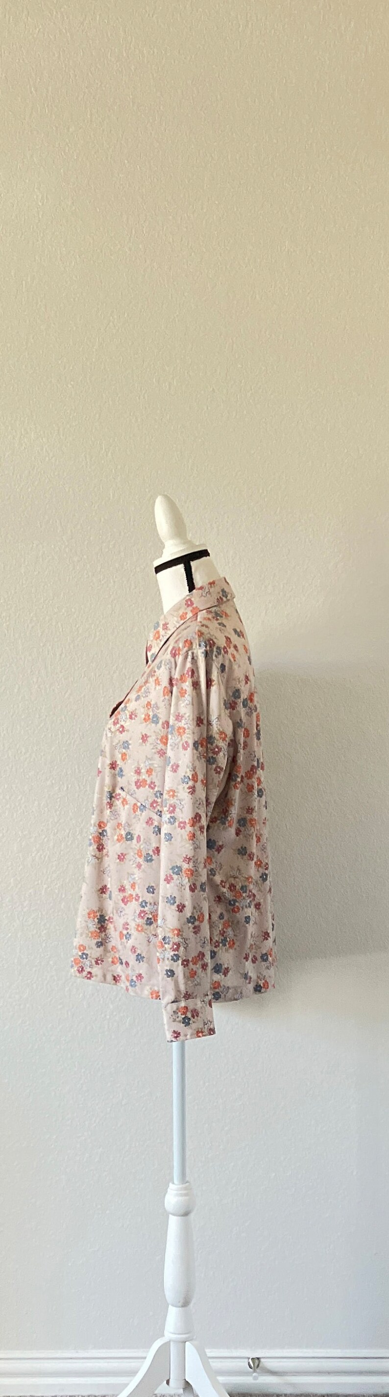 Puede incluir: Una camisa beige de manga larga con estampado floral de peque&ntilde;as flores rojas, azules y naranjas. La camisa est&aacute; en un maniqu&iacute; blanco con una banda negra en el cuello.
